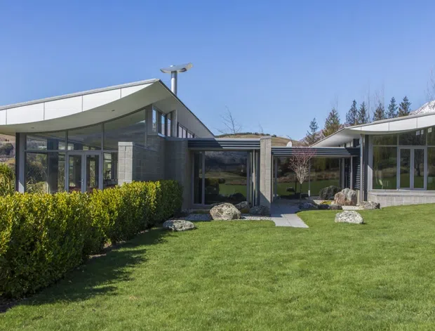 A low-profile contemporary single-storey residence set in a landscaped garden, with broad floor-to-ceiling glazing, separate curved roof forms, dark slender framing, and a central glazed connection. The property sits in an open, elevated landscape with hills visible beyond.