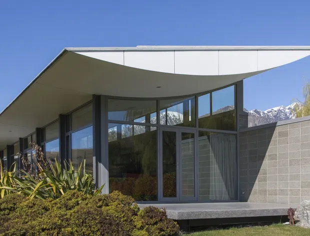 Single-storey modern house with expansive floor-to-ceiling glazing, a dramatic cantilevered flat roof with a curved underside, and a pale grey masonry feature wall. The building is set in landscaped grounds, with snow-capped mountains visible in the background and a clear blue sky.