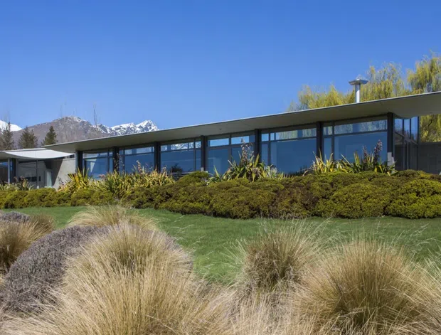 Low, elongated modern single-storey building set within a landscaped site, featuring extensive dark-framed glazing, a broad shallow roof with a pronounced overhang, and a sheltered entrance or side pavilion. Dense ornamental planting and grasses surround the building, with snow-capped mountains visible in the background.