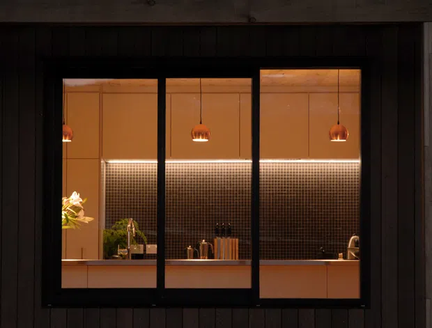 A dark exterior view through a large black-framed window into a warmly lit contemporary kitchen. The kitchen features pendant lights, a long illuminated small-format tiled splashback, timber-toned cabinetry, benchtops, and visible sink and tapware. The surrounding facade is clad in dark vertical timber boards.
