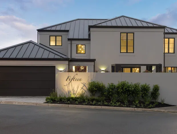 Street-facing view of a contemporary multi-storey residence with a separate double garage, dark standing-seam metal hipped roofs, smooth light-rendered walls, black-framed windows, and warm evening interior lighting. A low rendered boundary wall and landscaped planting screen the front entry; the facade has clean geometric massing and restrained modern detailing.