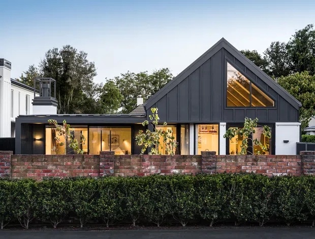 Street-facing exterior view of a contemporary single-storey home with a prominent dark charcoal gabled volume, vertical cladding, large glazed openings, and warm interior lighting. A lower white wing and chimney-like roof element sit to the left, while a low red-brown brick boundary wall and neatly clipped hedge define the front edge of the property.