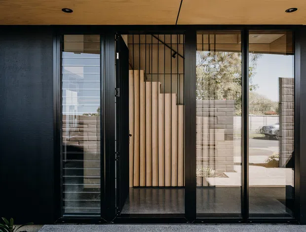 A contemporary residential entry facade viewed from outside, featuring a dark textured wall, black-framed glazed entry doors, and a timber-lined ceiling soffit. The central doorway opens to a compact entry hall with a striking vertical timber slat feature and black metal stair or balustrade detailing. Reflections and glazing reveal a paved suburban setting beyond.