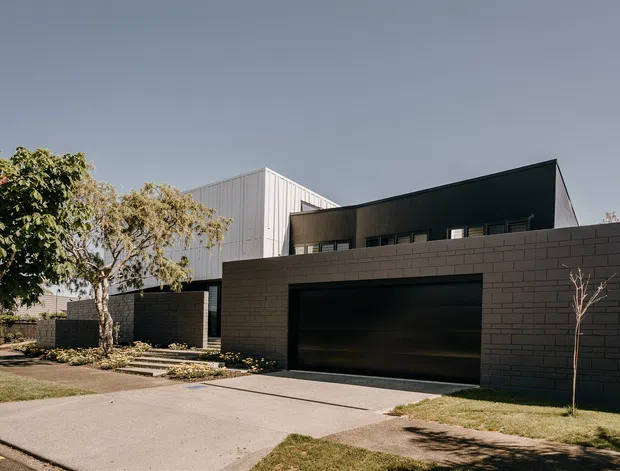 Exterior photograph of a contemporary multi-level residence viewed from the street, with a dark rendered or masonry garage frontage, broad integrated garage door, sharp angular roof forms, and a contrasting light vertical-clad upper volume. A paved driveway, low landscaping, and mature trees frame the facade.