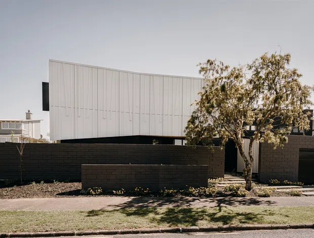 Exterior view of a contemporary, box-like residential building behind dark brick boundary walls. The upper volume is clad in pale vertically ribbed panels with a gently curved profile, paired with dark masonry walls, a recessed garage or entry opening, and a simple landscaped frontage with a mature tree.