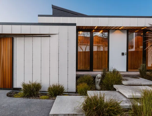 Exterior view of a contemporary single-storey residence with a low, angular roofline and crisp white panel cladding. The entry is framed by dark metal glazing, warm timber-lined soffits, and vertical timber screening, opening onto a landscaped path of large concrete stepping slabs surrounded by ornamental grasses.