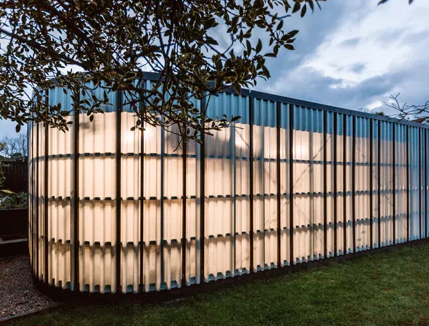 A compact modern building or pavilion viewed from outside at dusk, wrapped in translucent corrugated panels with prominent vertical dark framing members. Warm interior light glows through the translucent facade, while a darker blue corrugated upper band and flat roofline create a simple rectilinear form. The structure sits on a lawn and is partially screened by tree branches.