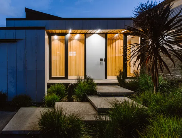 Dusk exterior view of a contemporary single-storey home entrance, featuring dark metal cladding, warm timber-lined recessed entry bays, broad glazing, concrete stepping slabs, and native-style landscaped planting. Strong contrast between the cool blue exterior light and warm illuminated entry.