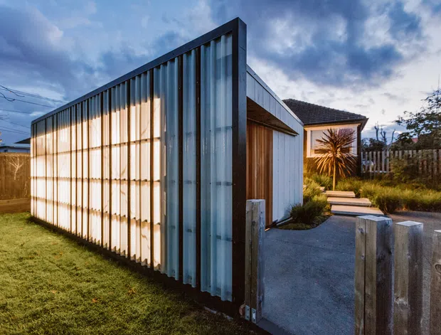 Exterior view of a compact contemporary residential building or extension with a strong vertical corrugated-metal screen facade, translucent glazing, timber cladding, and a simple dark pitched roof. A concrete path leads through a small landscaped front area, with timber fencing and low planting around the property. The image is photographed at dusk under a partly cloudy sky.