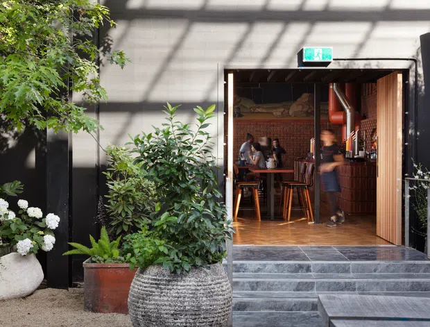 Exterior view of a dark contemporary commercial venue with a wide open timber-framed entrance, steps, and a warmly lit brick-lined interior visible inside. The entry is surrounded by lush planting, large potted greenery, hydrangeas, and textured stone planters. Interior details include timber flooring, bar-height stools, a counter, exposed brick, and visible ductwork.