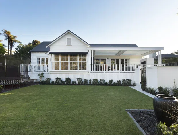 Exterior view of a white weatherboard-style single-storey house with a prominent gabled section, dark pitched roof, enclosed glazed veranda, white balustrades, and a covered side terrace. The home faces a neatly landscaped lawn with planting beds and a dark water feature or planter in the foreground.
