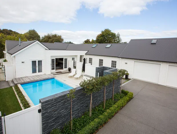 A single-storey residential property viewed from above, featuring white horizontal weatherboard cladding, a dark pitched metal roof with skylights, multiple garage doors, and a landscaped courtyard with a bright blue swimming pool, timber pool deck, and dark stone privacy wall.