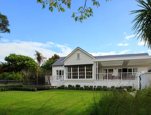 A single-storey suburban house viewed from the landscaped front yard. The home has a light weatherboard facade, a prominent gabled central section, a broad low-pitched roof extension, white-framed windows and doors, and a covered veranda with white balustrading. The property is surrounded by a manicured lawn, mature trees, ornamental grasses, and garden planting under a bright blue sky.