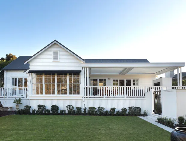 A bright exterior view of a single-storey white weatherboard home with a prominent front gable, dark pitched roof, enclosed veranda, white balustrades, and a covered carport or outdoor structure extending to the right. The property is set behind a neatly landscaped lawn with low hedging and a gated side path.