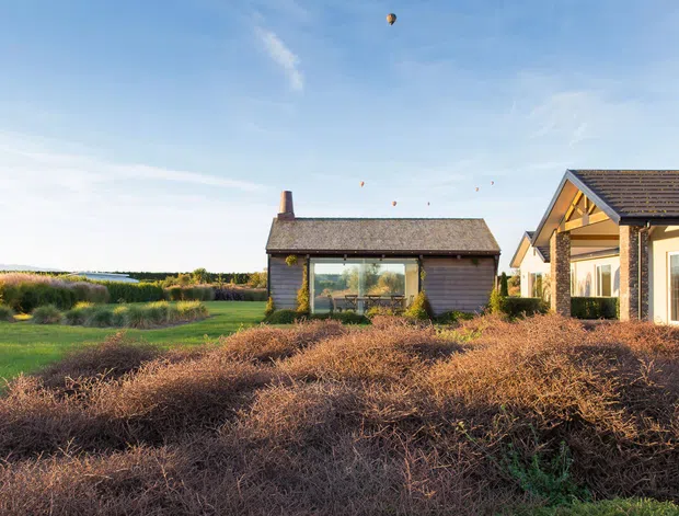 Exterior view of a low-rise rural property with a small dark-clad gabled outbuilding and an adjacent light-rendered building with a tiled pitched roof and exposed timber gable structure. The buildings sit in landscaped grounds overlooking open fields, photographed in bright daylight.