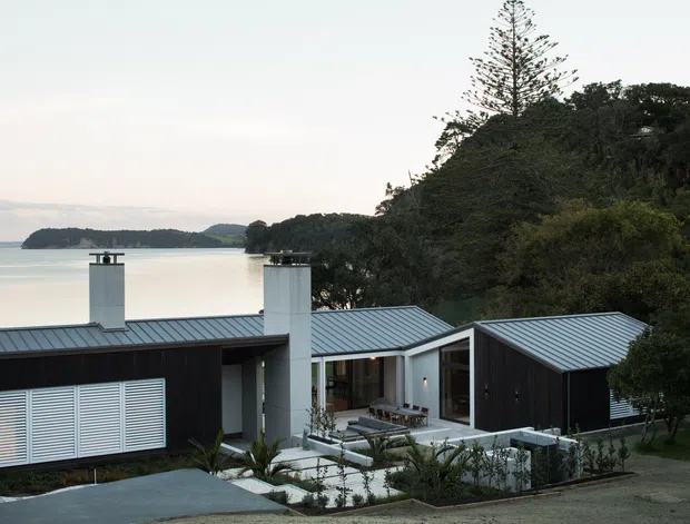 Exterior view of a contemporary low-rise residence with dark vertical cladding, pale metal pitched roofs, and two prominent white chimney volumes. The house is arranged around landscaped courtyards and outdoor seating, with a calm inlet or bay visible beyond the property and dense mature vegetation surrounding the site.