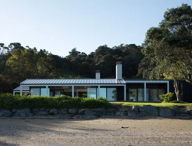 Single-storey contemporary residence viewed from the front, set amid dense mature vegetation. The home has a low horizontal profile, extensive floor-to-ceiling glazing, dark framed windows, pale metal roofing, and two simple chimney stacks. A gravel or sandy forecourt and low landscaped planting sit between the camera and the house.