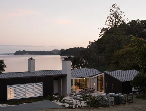 A modern single-storey waterfront residence viewed from an elevated exterior position. The home has dark vertical cladding, pale rendered chimney volumes, low-pitched metal roofing, expansive glazed openings and a landscaped central courtyard. A calm inlet and tree-covered shoreline are visible behind the property at dusk.
