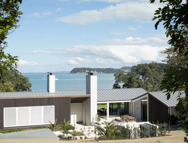 A low, modern coastal house viewed from above, with dark vertical timber cladding, a broad grey metal roof, two white chimney volumes, and large glazed openings around a sheltered outdoor courtyard. The house is set among trees on a sloping site overlooking bright blue water and a wooded headland. The architecture has a clean, contemporary pavilion-like composition with strong indoor-outdoor connection.