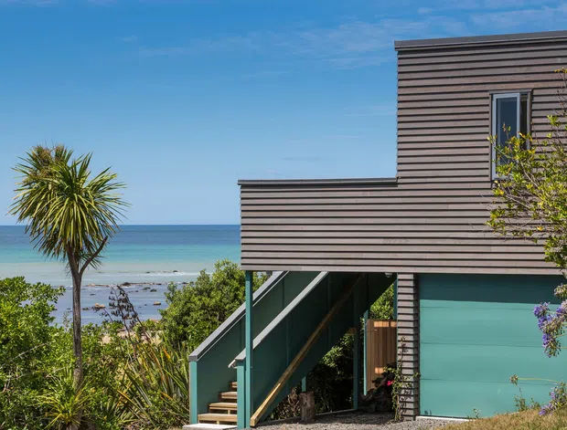 Exterior view of a contemporary multi-level coastal house overlooking the sea. The building has horizontally lined dark timber-toned cladding, a raised form with an open stairway beneath, turquoise painted lower walls, and simple rectangular windows. Coastal vegetation and a beach are visible around the property.