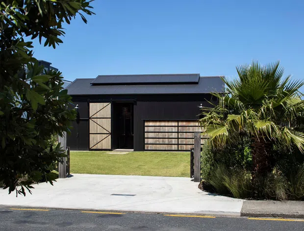 Street-facing exterior of a dark, single-storey contemporary building with a low-pitched metal roof, black vertical cladding, large timber-framed garage or barn-style doors, and a broad concrete driveway. Tropical and coastal planting partially screens the facade, with a manicured lawn and suburban street frontage.