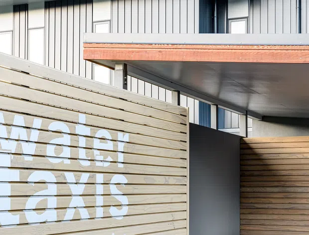 Close-up exterior architectural view of a contemporary building entrance or covered passage. The facade combines pale vertical metal cladding, dark framing, warm timber soffit and fascia detailing, and horizontal timber screening, including large white graphic lettering. A projecting flat roof or canopy dominates the upper right.