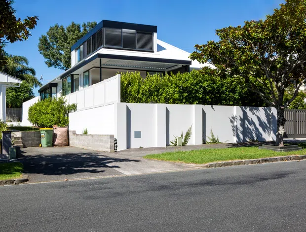 Street-facing exterior view of a contemporary multi-level residence with crisp white walls, extensive glazing, a projecting upper-level box with dark horizontal louvres, and a broad low-pitched roof. The property is enclosed by white privacy fencing and hedges, with a driveway and suburban street frontage.