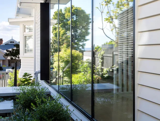 Exterior architectural photograph of a contemporary residential addition or facade, featuring a long wall of floor-to-ceiling glazing with slim dark frames, white horizontal weatherboard cladding, and a projecting soffit with recessed downlights. Dense landscaped planting runs along the base, while neighbouring residential buildings and greenery are reflected in the glass.