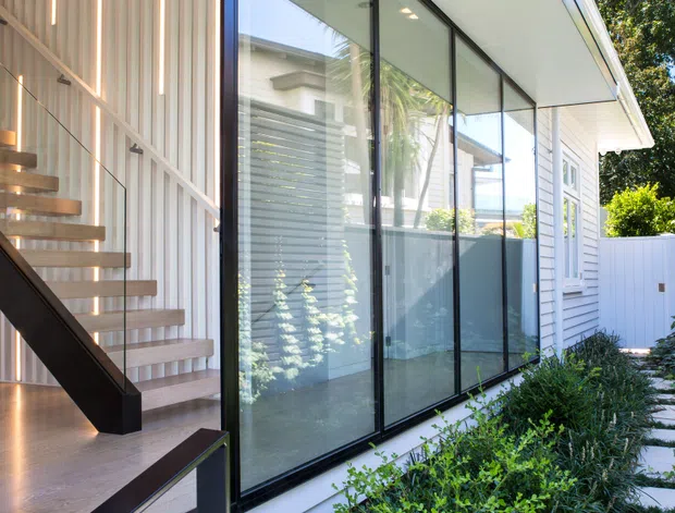 Exterior view of a contemporary residential entry and side courtyard. A glazed corridor or stair hall with full-height black-framed windows runs alongside a white weatherboard-clad house. The open stair has timber treads, a dark steel stringer and railing, while recessed ceiling lights sit beneath a broad white soffit. Pale concrete stepping slabs and dense low planting form the landscaped walkway.