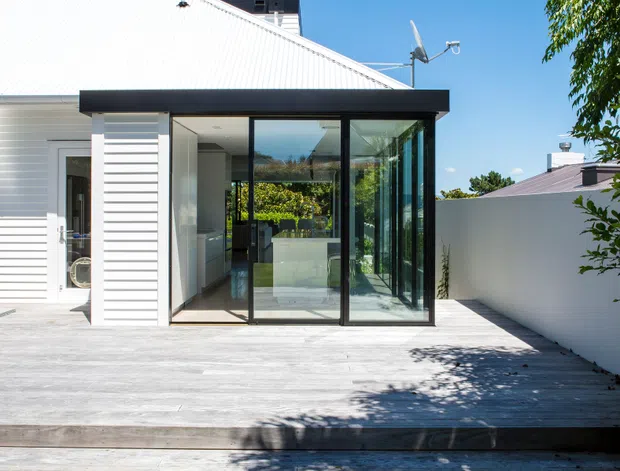 Rear exterior view of a light-coloured residential extension with a steep white corrugated metal gable roof, white horizontal cladding, and a black-framed glazed sunroom or living extension opening onto a broad timber deck. The deck steps down to a grass lawn, with a white boundary wall and neighbouring roof visible at the side. Bright clear daylight and strong shadows emphasise the crisp contemporary addition.