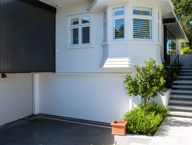 Bright exterior view of a renovated multi-level home, showing a white weatherboard facade with projecting bay windows, white metal pitched roof, horizontal window louvres, and a prominent integrated white garage door. A dark upper-level extension contrasts with the original facade. Steps, concrete paving, driveway, and neatly clipped hedge planting lead toward the entrance.