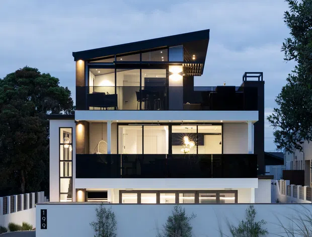 A dusk exterior view of a contemporary multi-storey residence with a strong horizontal composition, white rendered walls, dark charcoal or black balcony balustrades and window framing, and extensive glazing. The upper level has a dramatic sloping roofline and a covered outdoor terrace, while warm interior lighting is visible through the windows. A white boundary wall, landscaped planting, driveway, and neighbouring homes frame the property.