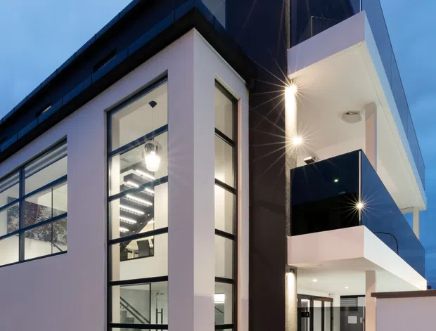 A dusk exterior view of a modern multi-storey building with crisp white rendered walls, black feature cladding, extensive black-framed floor-to-ceiling glazing, projecting balconies, and a covered ground-level entry or carport. Recessed exterior lighting creates strong starburst highlights, while a glimpse through the glazing shows a bright contemporary interior and stair. The composition emphasizes the building facade and vertical architectural form.