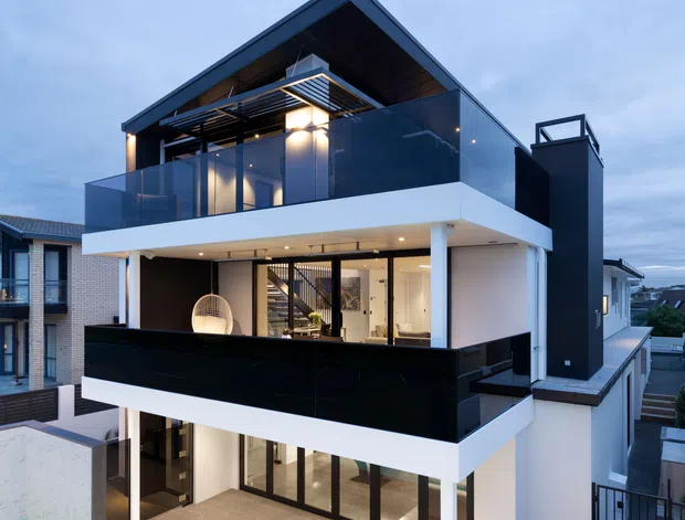 A multi-storey contemporary residence photographed from outside at dusk. The home features crisp white rendered volumes, dark charcoal cladding, extensive black-framed glazing, glass balcony balustrades, a projecting upper roof, and recessed exterior lighting. A narrow swimming pool and tiled outdoor dining area occupy the foreground, with neighbouring homes visible around the property.