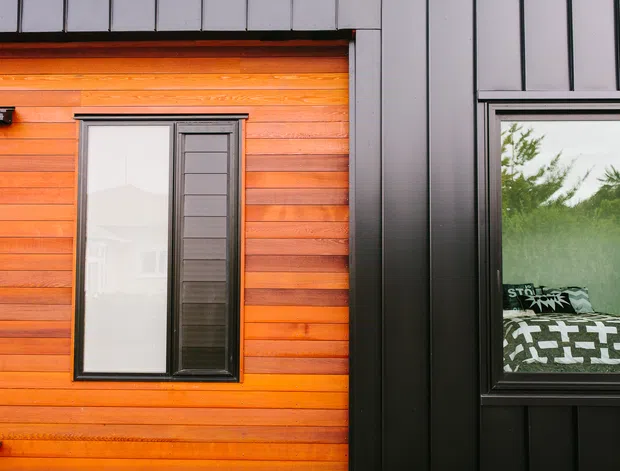 Close-up exterior view of a contemporary building facade combining warm horizontal timber cladding with dark vertical metal cladding and black-framed windows. The right window reveals part of an interior bedroom with patterned bedding, while the left window reflects the surrounding landscape.