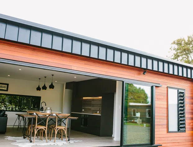 Exterior view of a contemporary single-storey extension or pavilion with warm horizontal timber cladding, dark metal roof edging, and large glazed openings. The open facade reveals a combined kitchen and dining area with a timber dining table, pendant lights, dark cabinetry, and indoor-outdoor connection.