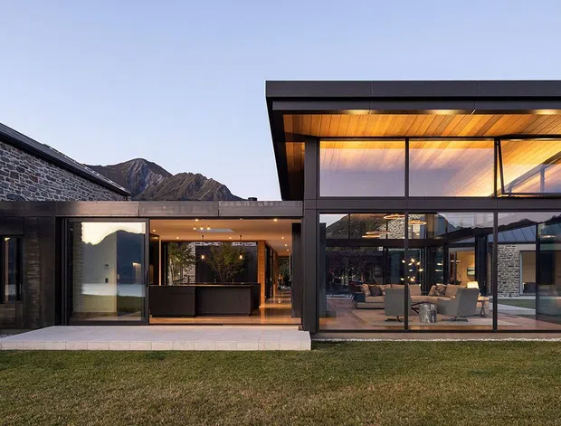 Exterior architectural view of a contemporary multi-wing residence set against a mountainous landscape. The home combines dark masonry walls, extensive floor-to-ceiling glazing, strong black structural framing, and a prominent flat-roofed double-height pavilion with warm timber soffit lining. A lawn occupies the foreground, while furnished interior spaces are visible through the glazing.