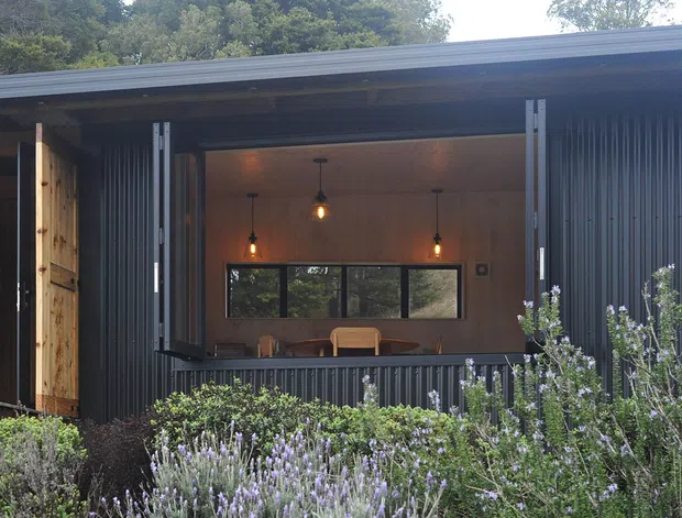 Exterior view of a compact, single-storey building with dark vertically corrugated cladding and a broad open folding window or servery. Warm timber-lined interior walls, pendant lights, and a dining table are visible through the opening. Dense landscaped planting occupies the foreground, and the low roof has a simple modern profile.