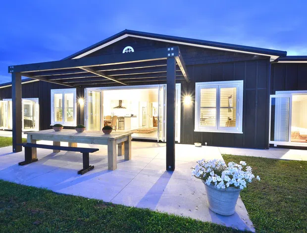Exterior view of a dark-clad single-storey home at dusk, featuring a covered outdoor entertaining area with a timber dining table and bench, paved terrace, illuminated interiors visible through open doors and windows, and a simple pitched roof.