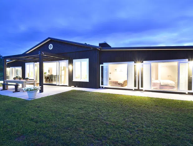 Exterior dusk view of a long, single-storey dark-clad home with a pitched gable section and broad low roofline. The facade has extensive illuminated glazing, projecting window bays, a covered outdoor entertaining area with dining furniture, and a lawned garden setting.