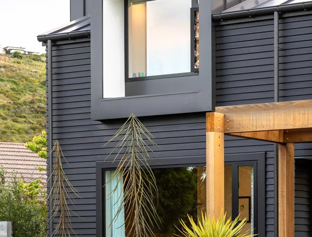 Exterior view of a contemporary multi-level home with dark horizontal cladding, a projecting box window, metal roof elements, and a timber-framed covered entry or outdoor structure. The landscaped foreground includes clipped hedges and architectural planting, while a sloping hillside is visible in the background.