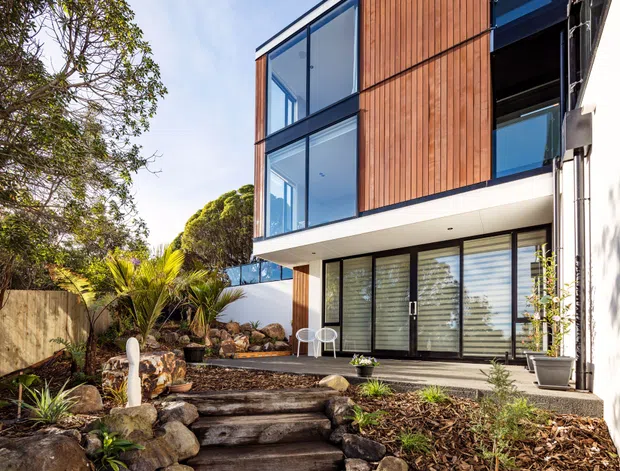 Exterior view of a contemporary multi-storey home set within a landscaped garden. The facade combines warm vertical timber cladding, extensive black-framed glazing, white rendered projecting volumes, and a dark roof edge. Timber-log garden steps, natural stone retaining edges, rocks, and subtropical planting lead toward a glazed ground-level living area and upper-floor rooms.