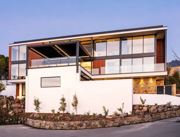 A contemporary multi-storey residence viewed from the street at dusk. The home features a flat roof, extensive floor-to-ceiling glazing, dark framed windows, warm timber-lined balcony and soffit areas, white rendered walls, and a prominent natural stone wall. Landscaped planting and low stone retaining walls frame the facade.