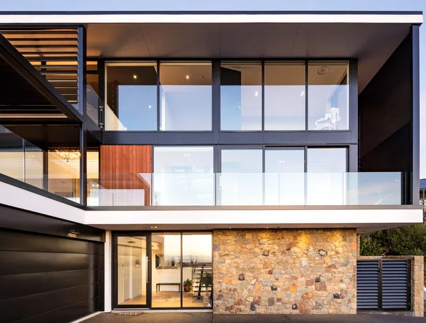Exterior architectural photograph of a contemporary multi-level residence with a strong rectilinear form, flat roof, extensive floor-to-ceiling glazing, dark framed windows, glass balcony balustrades, timber screening, a dark garage door, and a prominent natural stone feature wall at ground level.