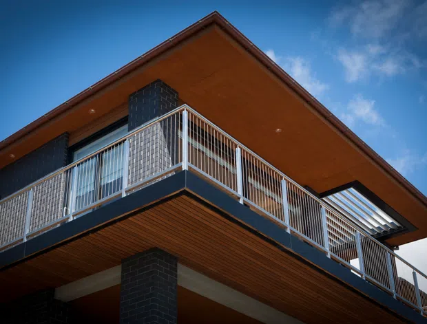 Low-angle exterior view of a contemporary multi-level building with broad projecting eaves, warm timber soffits, dark brick masonry, and a wraparound balcony enclosed by slim white metal railings. A roof or upper-level skylight is visible against a blue sky.