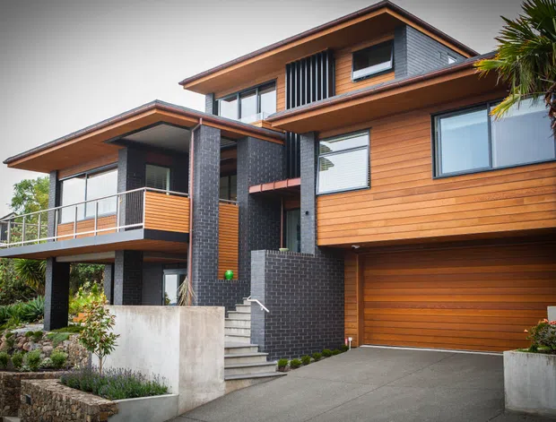 Exterior view of a contemporary multi-level residence on a sloped site, featuring warm horizontal timber cladding, dark brick masonry, broad roof overhangs, large glazed windows, a covered balcony, and an integrated double garage. Landscaping and a palm tree frame the driveway and entry stairs.