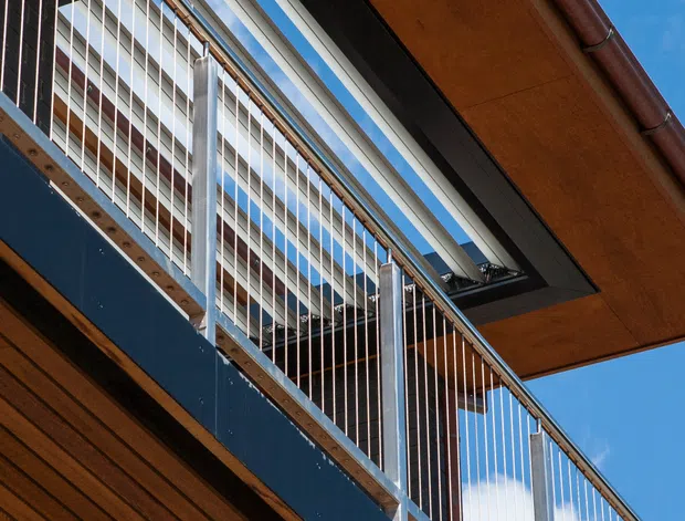 Upward-looking architectural detail of a contemporary multi-level exterior, featuring warm timber soffits and cladding, dark blue fascia panels, slender stainless-steel cable balustrades, metal framing, and a large roof or canopy opening with visible sky beyond. The image emphasizes layered decks, projecting roof planes, and crisp structural geometry against a bright blue sky.