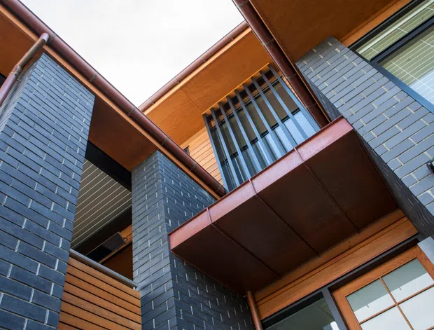 Upward-looking view of a multi-level contemporary building facade arranged around an open courtyard or void. Dark blue-grey brick piers contrast with warm timber soffits, horizontal timber cladding, reddish-brown metal roof edges and balcony undersides, and large glazed openings. Vertical metal screens and projecting balcony forms create a strong geometric composition.