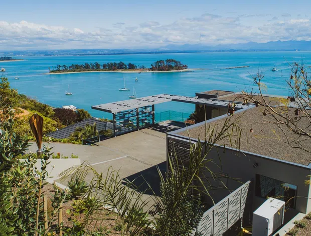 A wide exterior view of a contemporary waterfront property overlooking a vivid turquoise bay. Flat-roofed dark and light building volumes, a broad paved terrace, and a covered outdoor structure sit amid landscaped coastal vegetation, with small islands and distant mountains across the water.