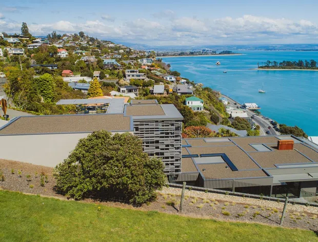 Aerial view of a contemporary coastal residential property on a sloping site, with broad low-pitched roof forms, extensive glazing and horizontal architectural detailing. The house overlooks a bright blue sheltered bay with boats, while surrounding hillside homes, mature vegetation and landscaped grounds establish a suburban waterfront setting.